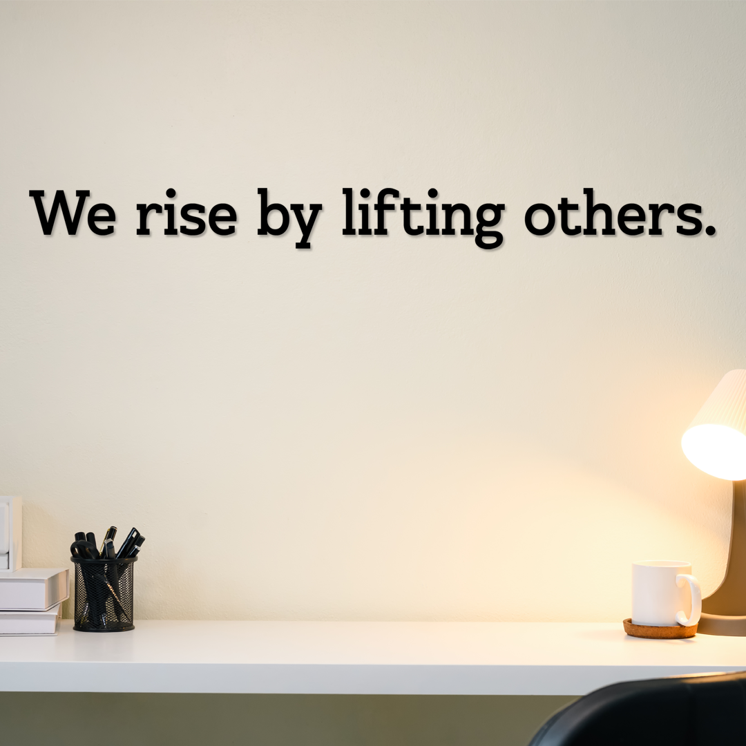 Motivational quote 'We rise by lifting others.' on a wall above a desk with office supplies. Acrylic wall decor - Vibewall