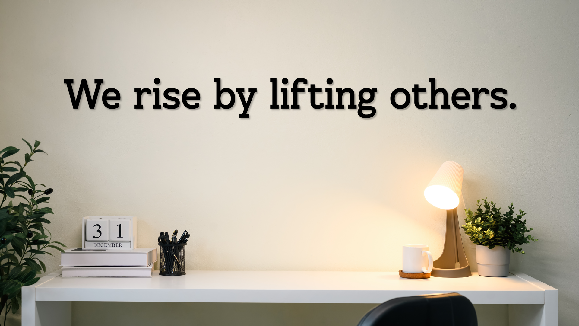 Decorative wall quote 'We rise by lifting others.' in a home office setting with a desk and plants. Acrylic wall decor - Vibewall