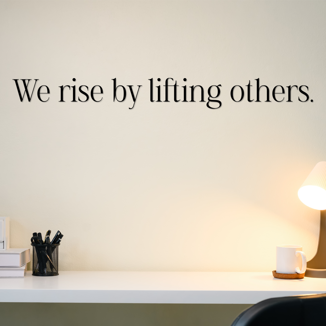 Wall with motivational quote 'We rise by lifting others' above a desk with office supplies. Acrylic wall decor - Vibewall