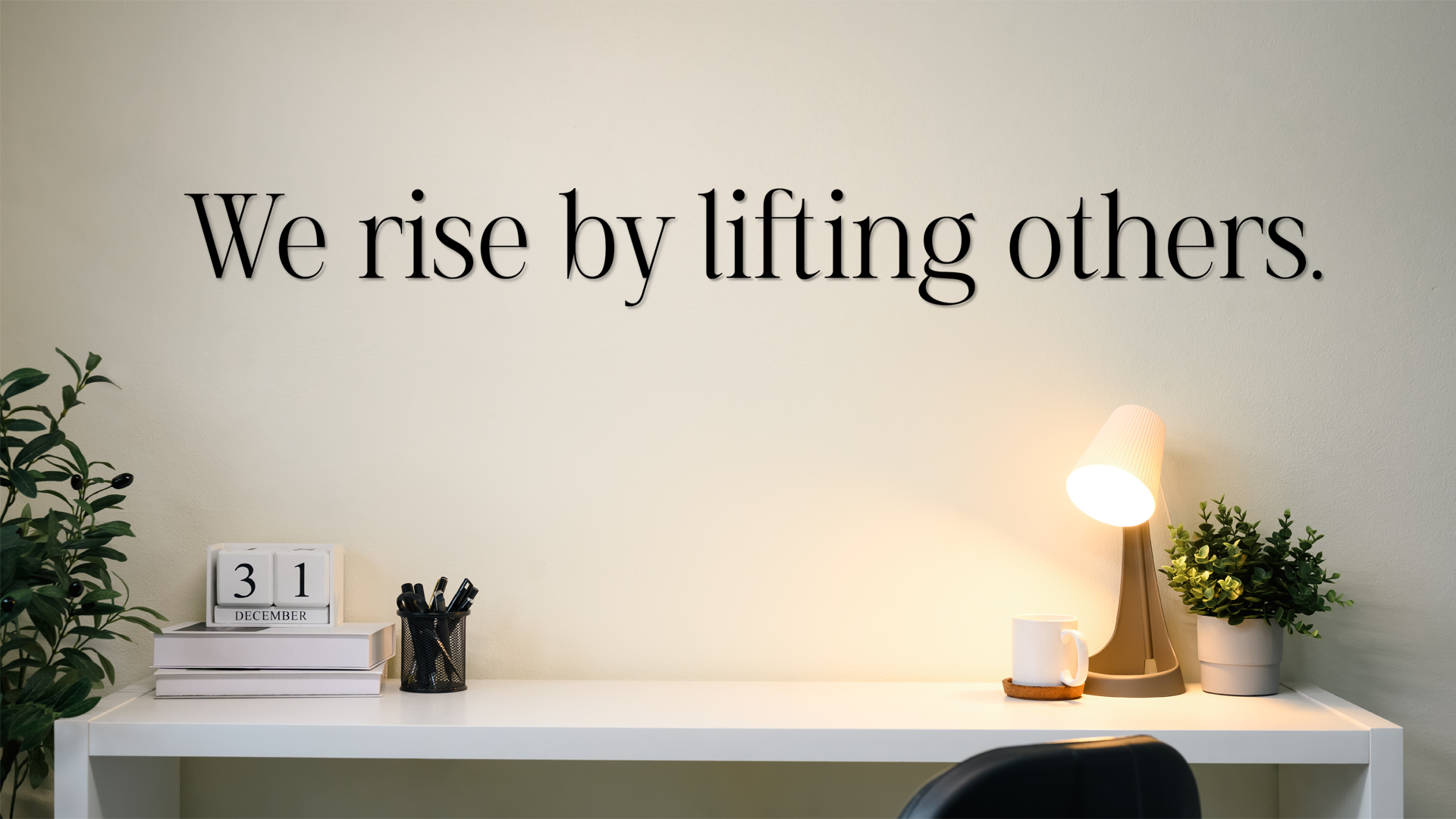 Decorative wall quote 'We rise by lifting others' in a home office setting with a desk and plants. Acrylic wall decor - Vibewall