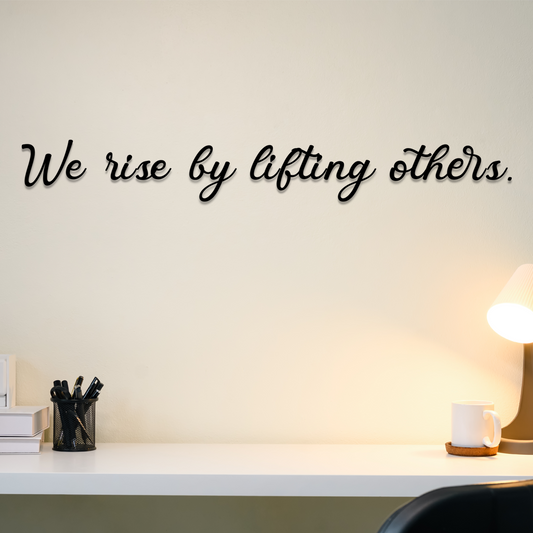 Wall with motivational quote 'We rise by lifting others' above a desk with office supplies. Acrylic wall decor - Vibewall